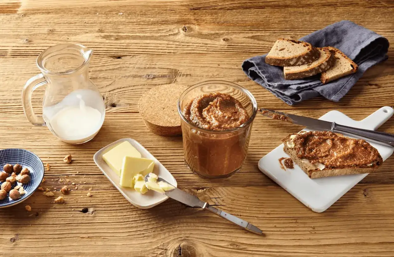 Zutaten f&uuml;r nougat selber machen: Haseln&uuml;sse, Butter, Milch und Brot. Ein Glas mit selbstgemachtem Nougat steht bereit.
