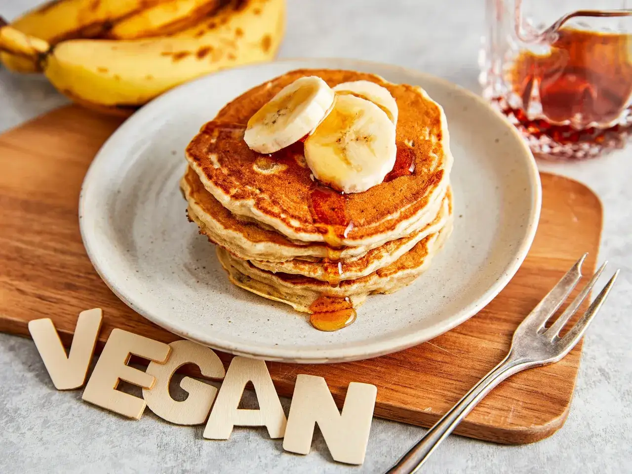 Stapel fluffiger, goldbrauner bananen pancakes ohne ei, garniert mit Bananenscheiben und Ahornsirup. Daneben liegen Bananen und ein Gabel.