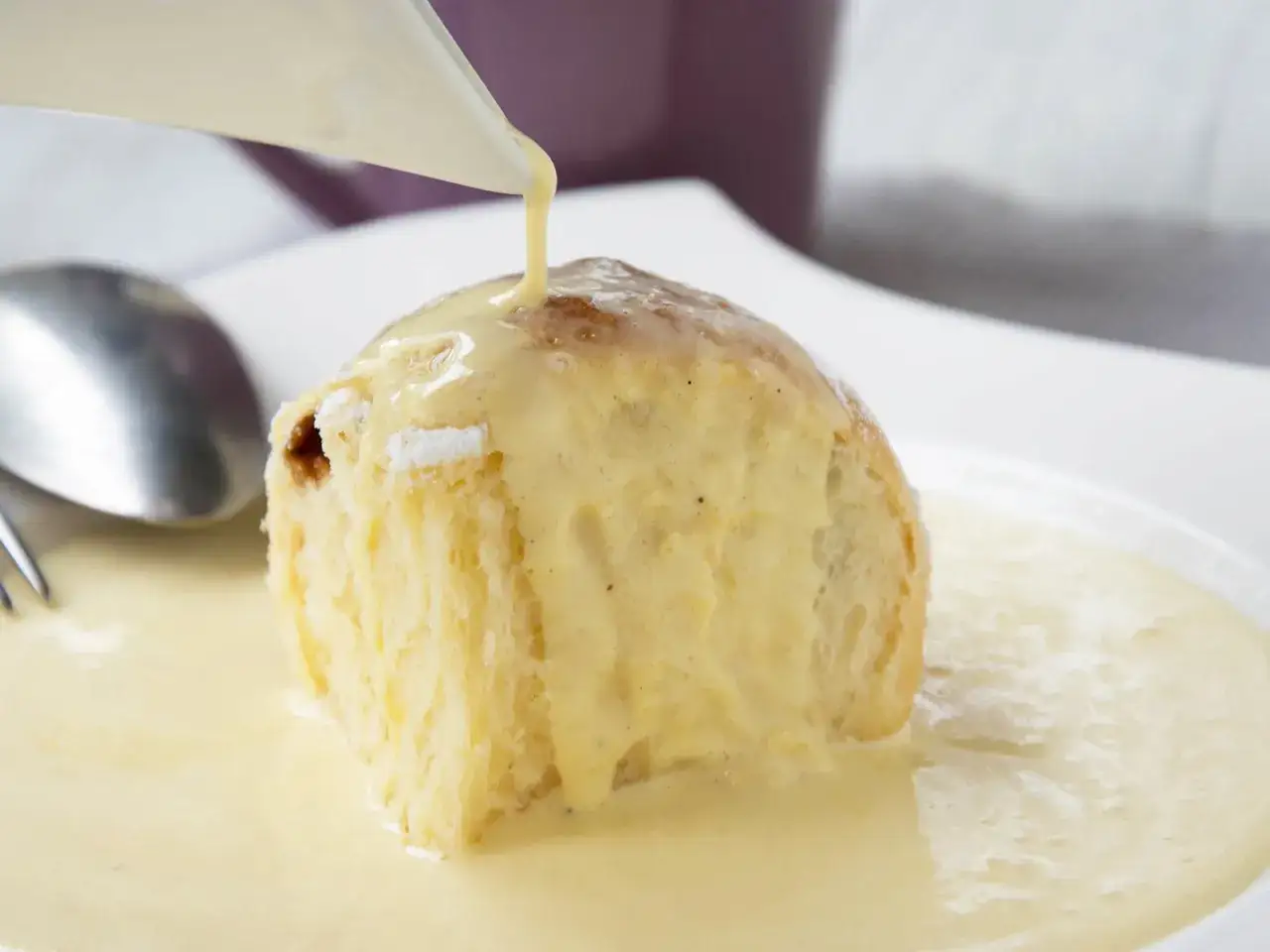 Ein flaumiger Germkn&ouml;del wird mit selbstgemachter Vanillesauce &uuml;bergossen. Ein L&ouml;ffel liegt daneben.