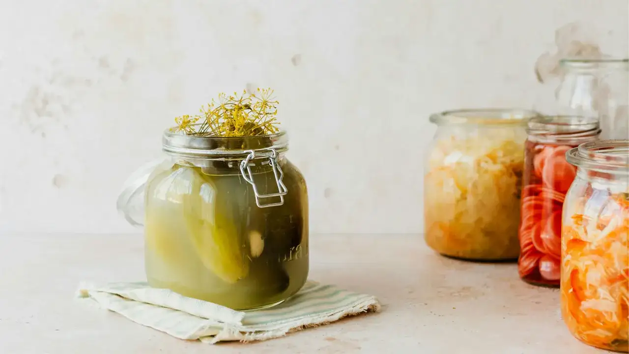 Ein Glas mit Dill und Gurken, bereit, um Salzgurken selber machen. Daneben stehen weitere Gl&auml;ser mit fermentiertem Gem&uuml;se.