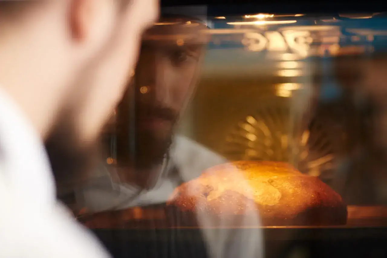 Ein Mann mit Bart schaut in den Ofen, wo ein Brot b&auml;ckt. F&uuml;r knuspriges Brot muss man beim Brot backen Wasser in den Ofen.