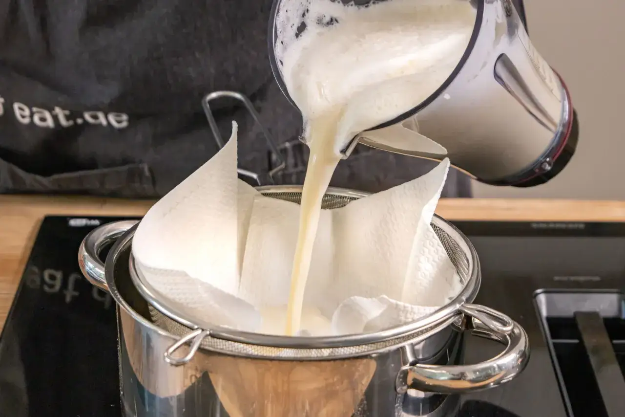 Fl&uuml;ssigkeit aus einem Mixer wird durch ein Sieb mit K&uuml;chenpapier gegossen, um sojamilch selber machen zu erleichtern.